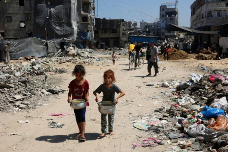 Des enfants palestiniens portent au milieu des destructions et des déchets, des marmites de soupe reçue à un point de distribution de nourriture dans le camp de réfugiés de Jabalia, dans le nord de la bande de Gaza, le 21 août 2024 ( AFP / Omar AL-QATTAA )