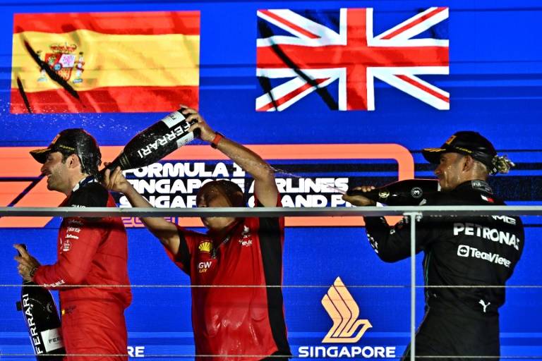 Le patron de l'écurie Ferrari Frederic Vasseur (au centre) et le pilote Mercedes Lewis Hamilton(à droite) arrosent de vin mousseux Carlos Sainz (à gauche), vainqueur du Grand Prix de Singapour 2023, sur le circuit de Marina Bay le 17 septembre 2023. ( AFP / Lillian SUWANRUMPHA )
