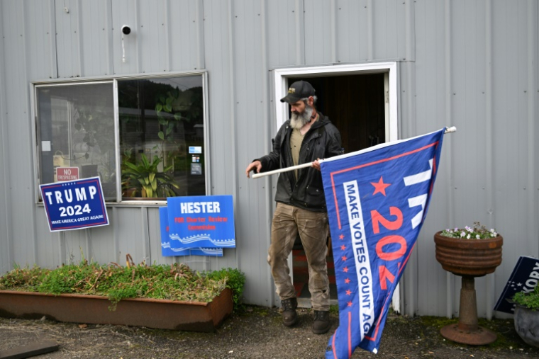 Hickory Grant, membre du conseil municipal de Forks, tient un drapeau "Trump 2024" devant son atelier de réparation, le 15 octobre 2024 dans le comté de Clallam de l'Etat de Washington ( AFP / Robyn Beck )