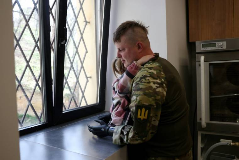 Le sergent ukrainien Valeriï Koutcherenko, équipé de prothèses bioniques, tient dans ses bras sa plus jeune fille, Oleksandra, dans leur appartement à Bila Tserkva, au sud de Kiev, le 2 avril 2024 ( AFP / Anatolii STEPANOV )
