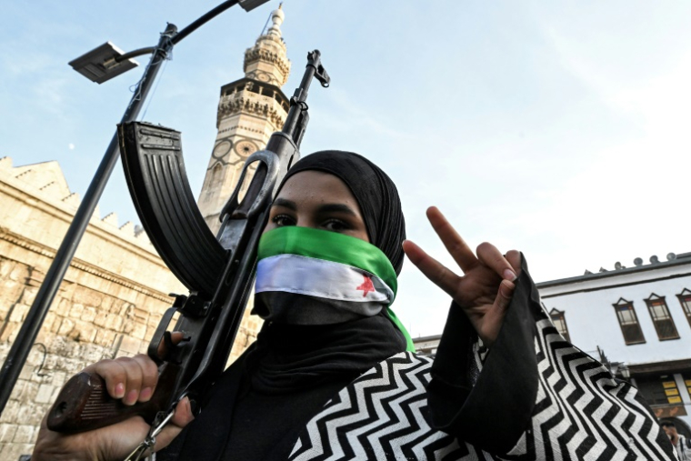 Une femme masquant son visage avec le drapeau de l'opposition syrienne fait le V de la victoire en tenant un fusil d'assaut devant la mosquée des Omeyyades à Damas le 10 décembre 2024 ( AFP / LOUAI BESHARA  )